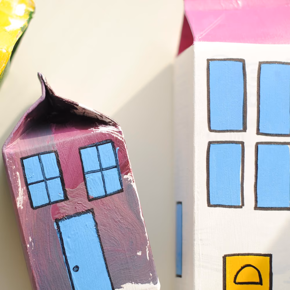 Zwei kleine Häuser aus bemalten Milchkartons; links ein lila Haus mit blauem Fenster und Tür, rechts ein weißes Haus mit vier blauen Fenstern und einer gelben Tür.
