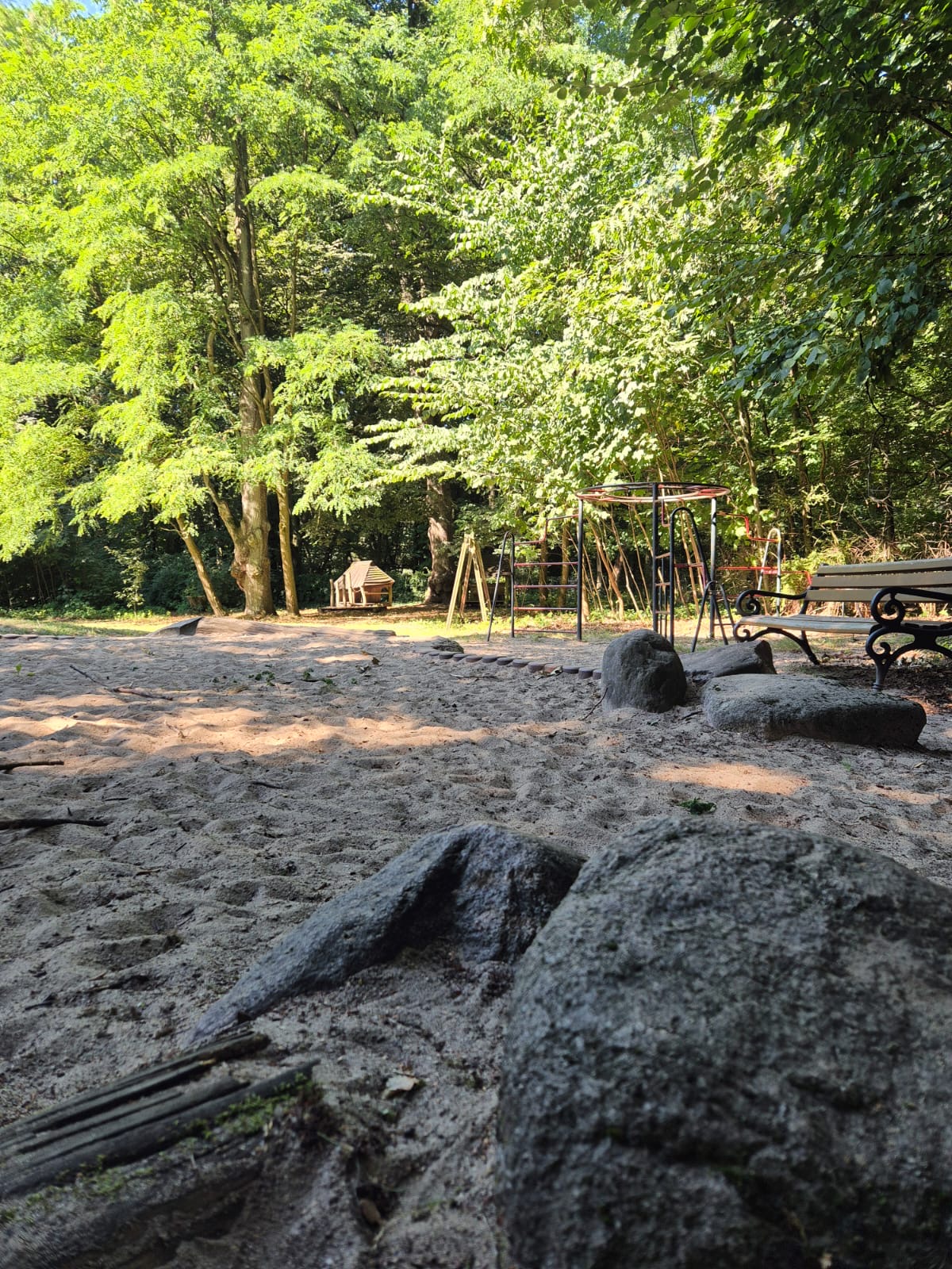 Spielplatz mit Sandfläche, Klettergerüst, Schaukel und einer Bank, umgeben von dichtem, grünen Laub und Bäumen an einem sonnigen Tag.