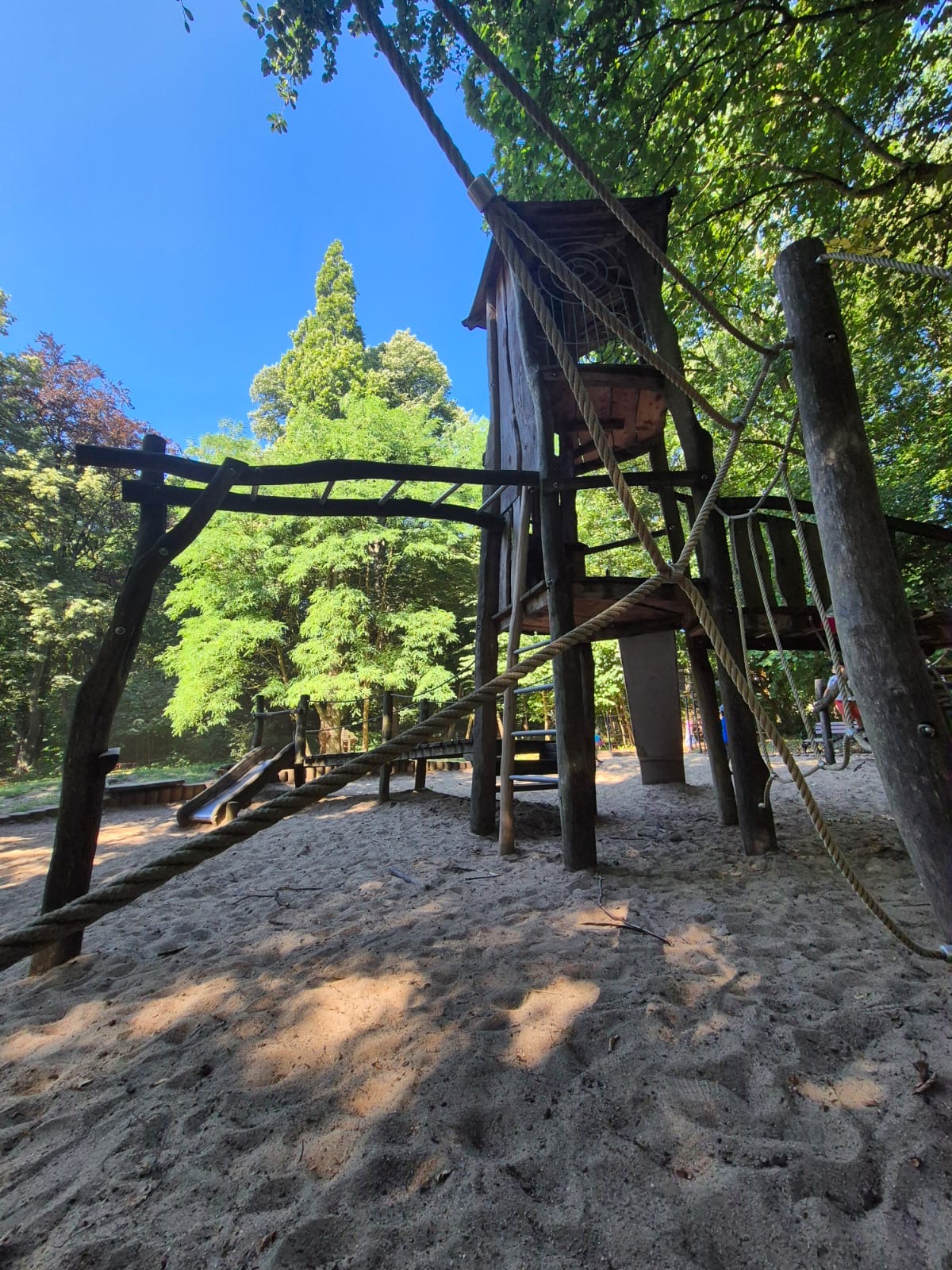 Kinderspielplatz mit Holzklettergerüst, Rutsche, Seilen und Sandboden, umgeben von grünen Bäumen unter blauem Himmel.