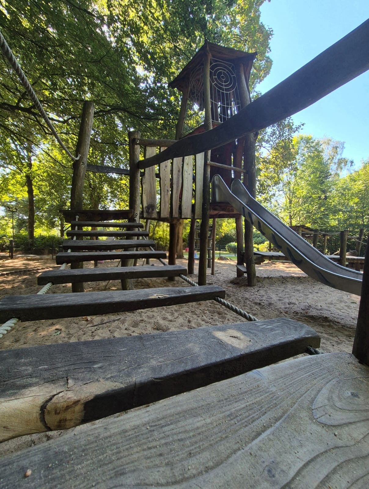 Spielplatz im Freien mit einer Holzkletterstruktur, die eine Rutsche und Balancierbalken über Sandboden enthält, umgeben von grünen Bäumen.