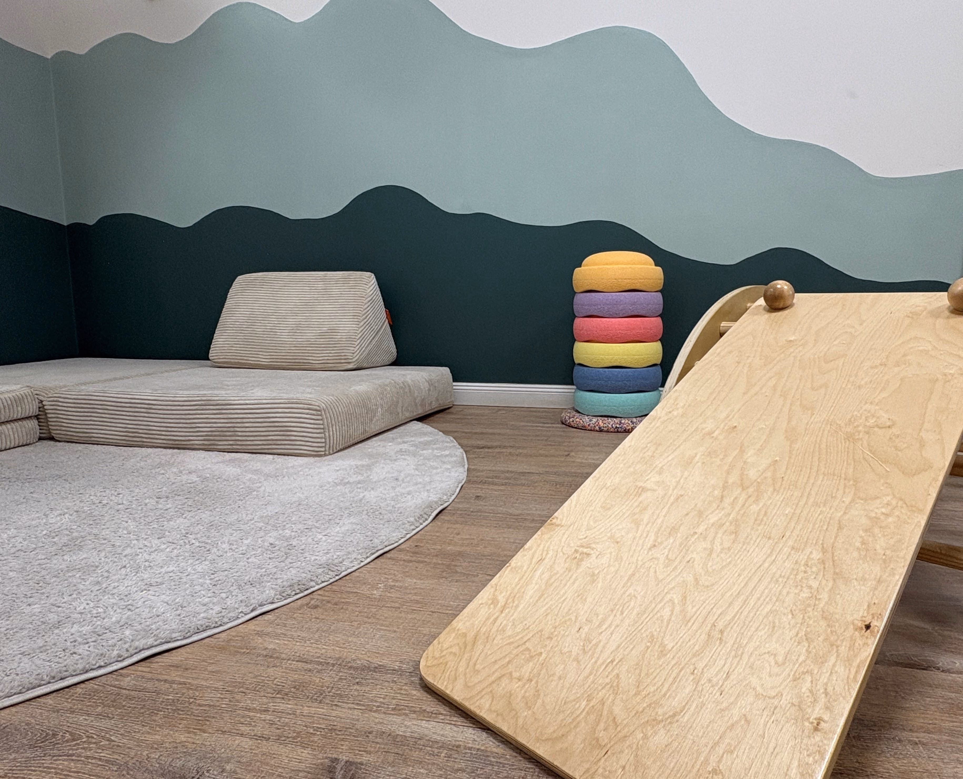 Kinderspielecke mit einem weichen, grauen Teppich, einer gepolsterten, hellbeigen Sitzmatte an der Wand und einer hölzernen Rutsche mit bunten Polsterkissen im Hintergrund vor einer mehrfarbigen Wand mit wellenförmigem Design.