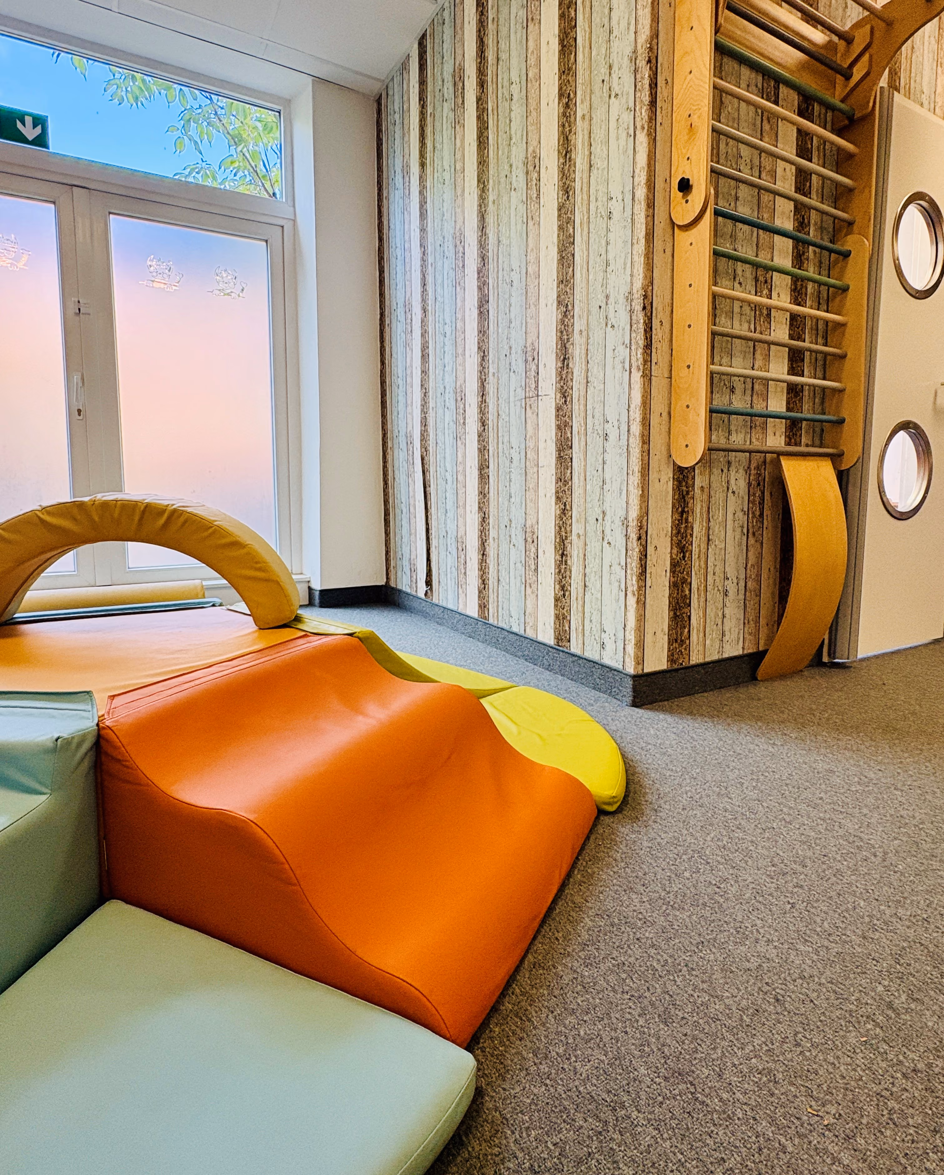 Helle Kinderspielecke mit bunten weichen Kletterelementen in Orange, Gelb und Grün, Holzleiter an der Wand und einem Fenster mit halbdurchsichtigen Vorhängen.