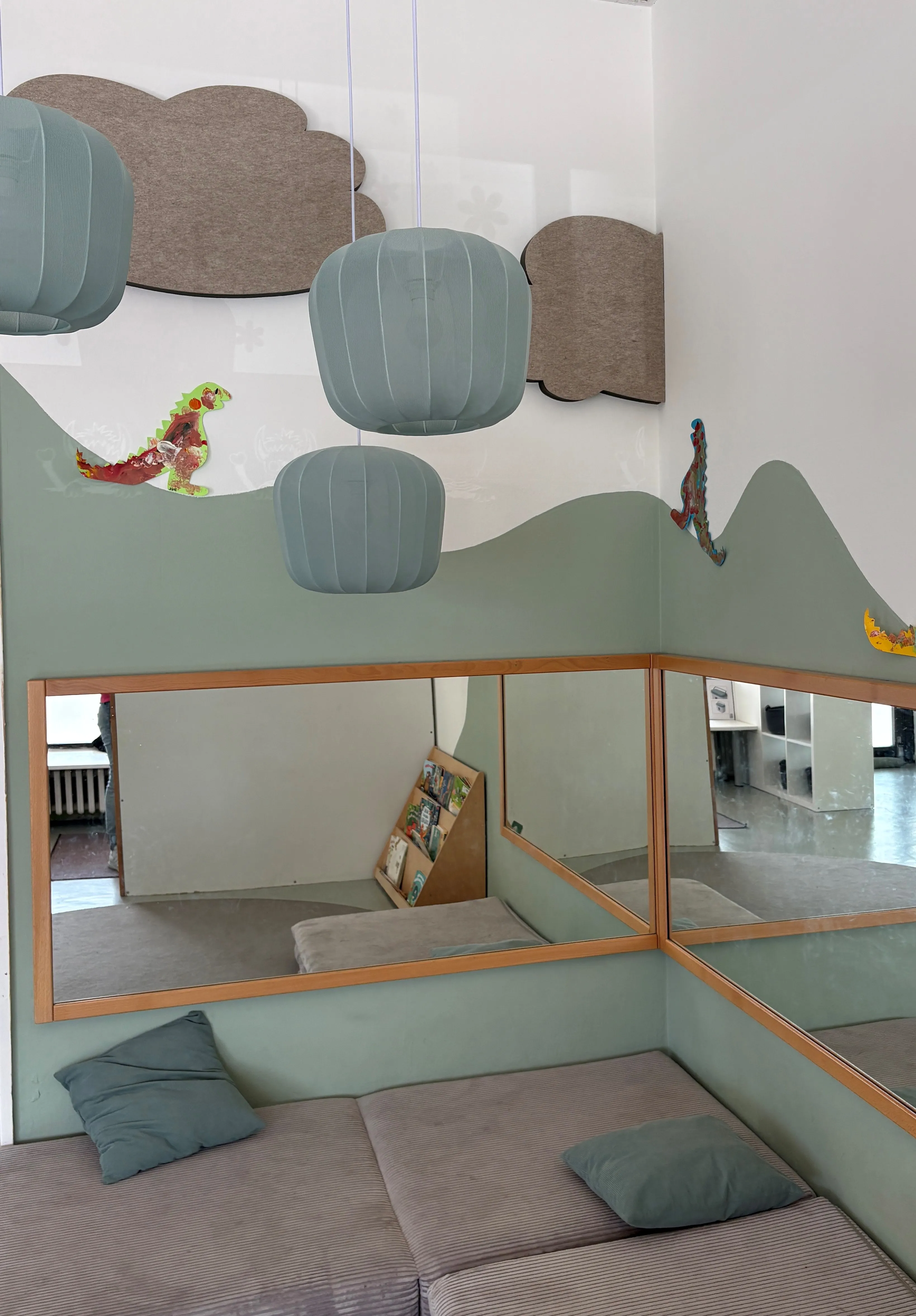 Gemütliche Ecke mit gepolstertem Sitzbereich, zwei Kissen und großen Spiegeln an den Wänden, darüber hängend drei blaue Lampen und dekorative Dinosaurierfiguren auf hellgrünem Hintergrund mit wolkenförmigen Akzenten.
