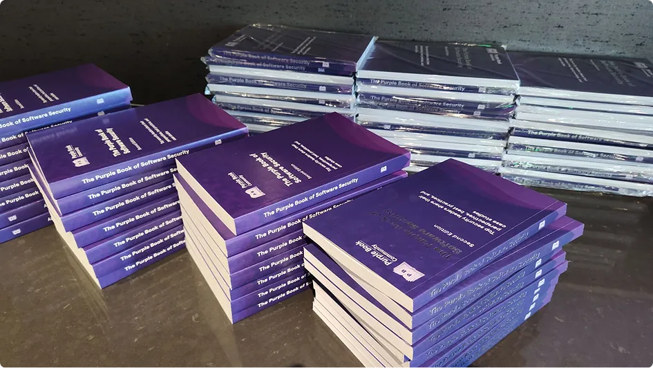Stacks of the Purple Book of Software Security in hardcover and paperback on a table.