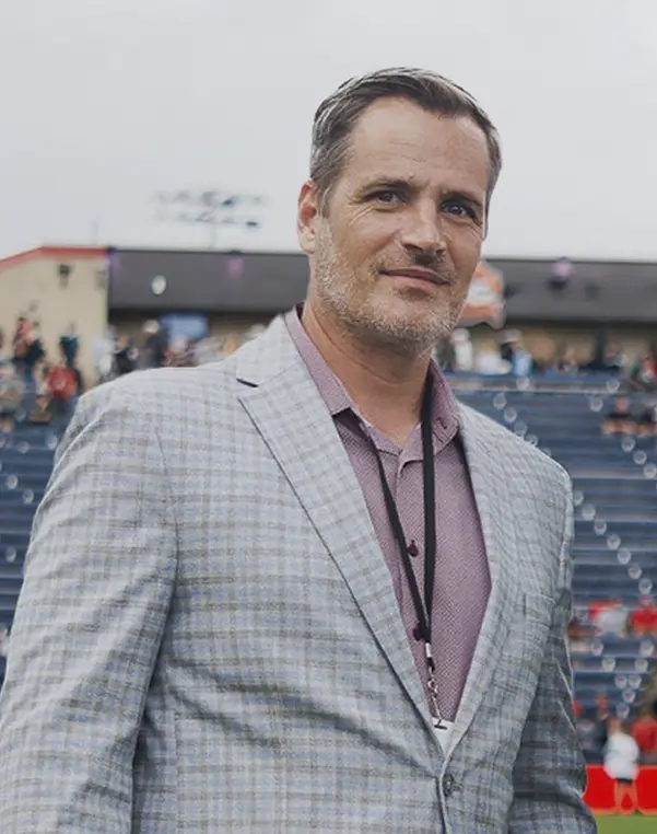 Man wearing a light gray plaid blazer and a maroon shirt standing outdoors with stadium seats and people blurred in the background.