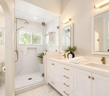 White bathroom with brass faucets and light fixtures, a double vanity and glass shower