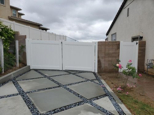 Driveway with geometric concrete slabs separated by dark pebbles leading to white double gates with a small white side gate next to a brown brick wall and a rose bush on the right.