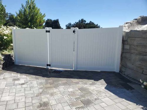 White double gate with black hinges on a paved driveway, bordered by green trees and stone walls under a clear blue sky.