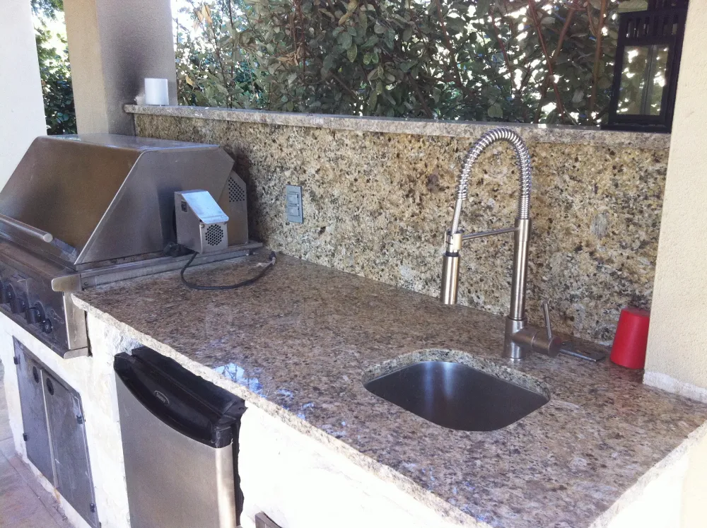  Outdoor kitchen with speckled brown granite countertops, stainless steel grill, sink, and mini fridge.
