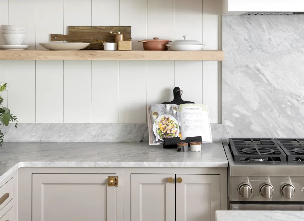 Bright kitchen with white marble countertops and backsplash, light gray cabinets, and wooden floating shelf.