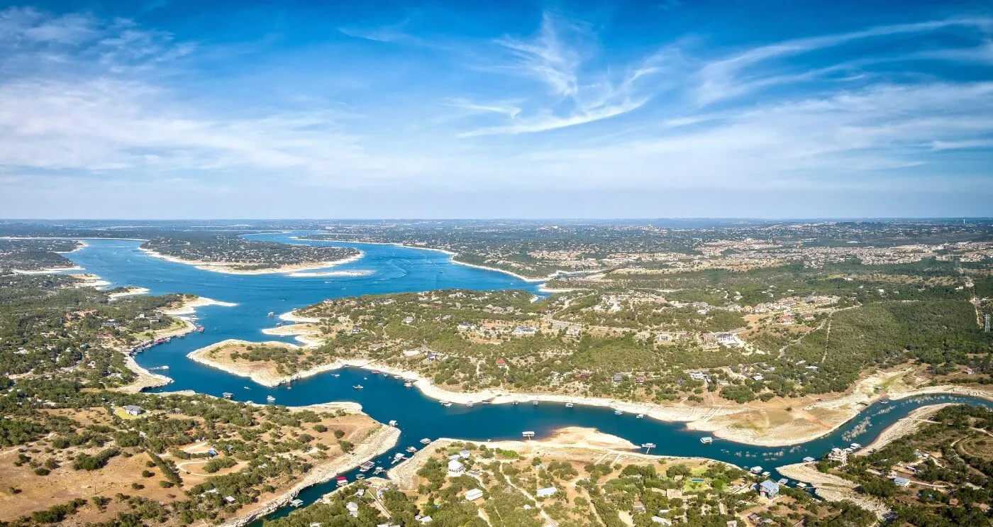 Aerial view of a winding lake surrounded by hills, homes, and vegetation under a bright blue sky.