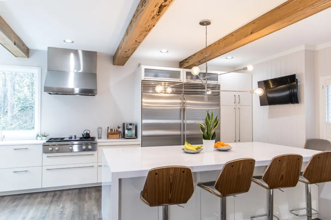Modern white kitchen with exposed wooden beams, stainless steel appliances, and a white waterfall island with wooden barstools.