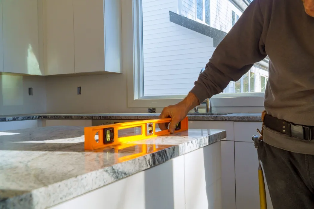 Person using a level tool to check the evenness of a newly installed granite countertop in a bright kitchen with white cabinets and large window.