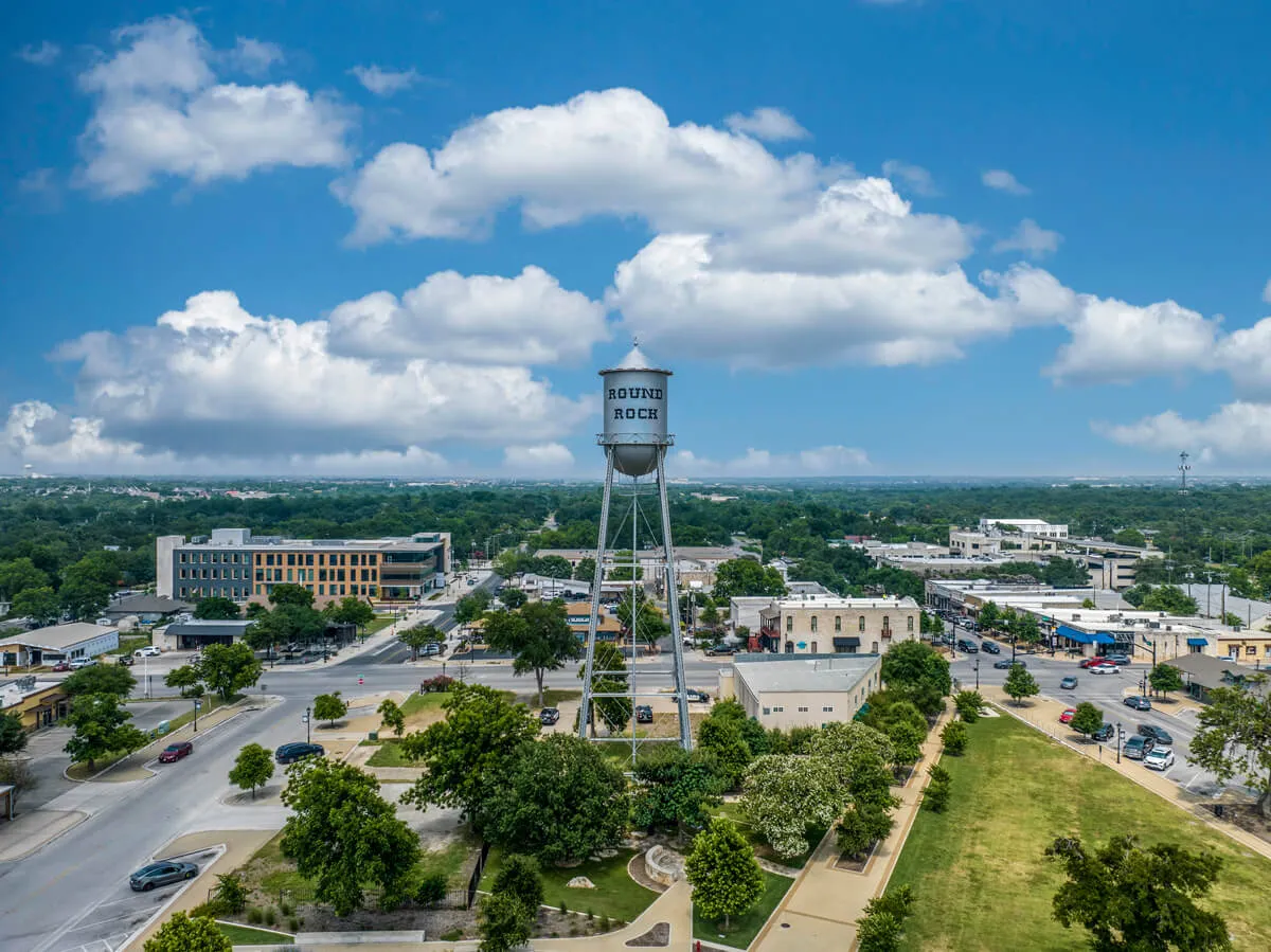 Aerial view of downtown Round Rock, Texas, featuring the historic water tower surrounded by buildings, trees, roads, and a partly cloudy sky.