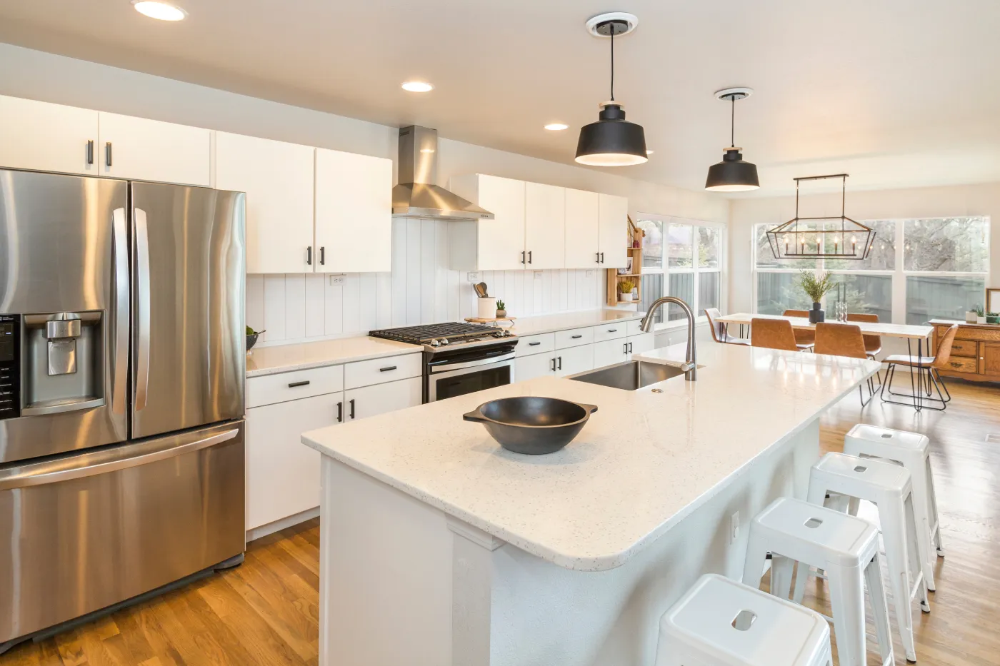Modern kitchen with white cabinetry, stainless steel appliances, a large island with a light quartz countertop, black pendant lights, and an adjacent dining area with large windows.