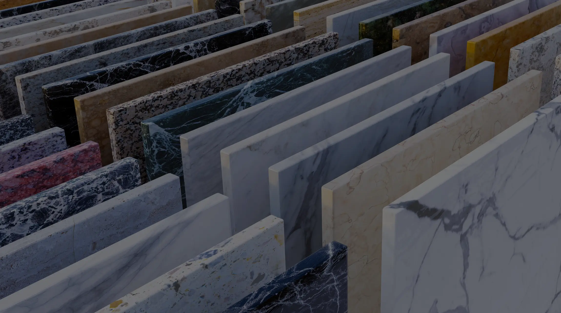 A collection of large, polished marble and granite slabs in various colors and patterns, including white, black, beige, green, and red, neatly arranged in parallel rows at a stone warehouse. The surfaces reflect light, highlighting the natural veining and textures of each piece.