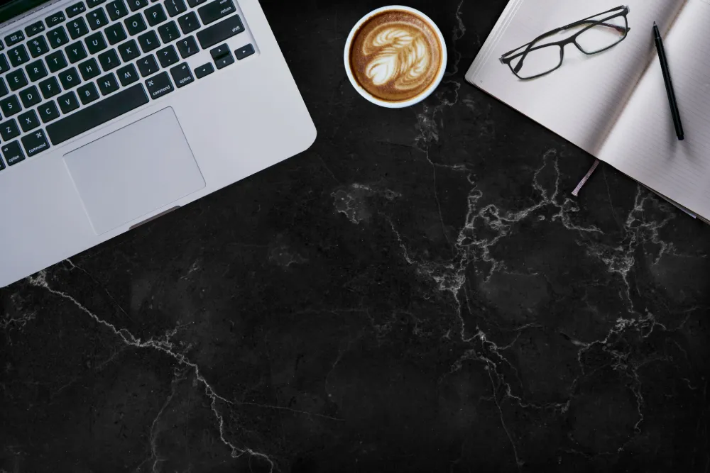 Top view of a workspace with a laptop, a cup of latte art coffee, an open notebook, black-framed glasses, and a pen on a black quartz desk.