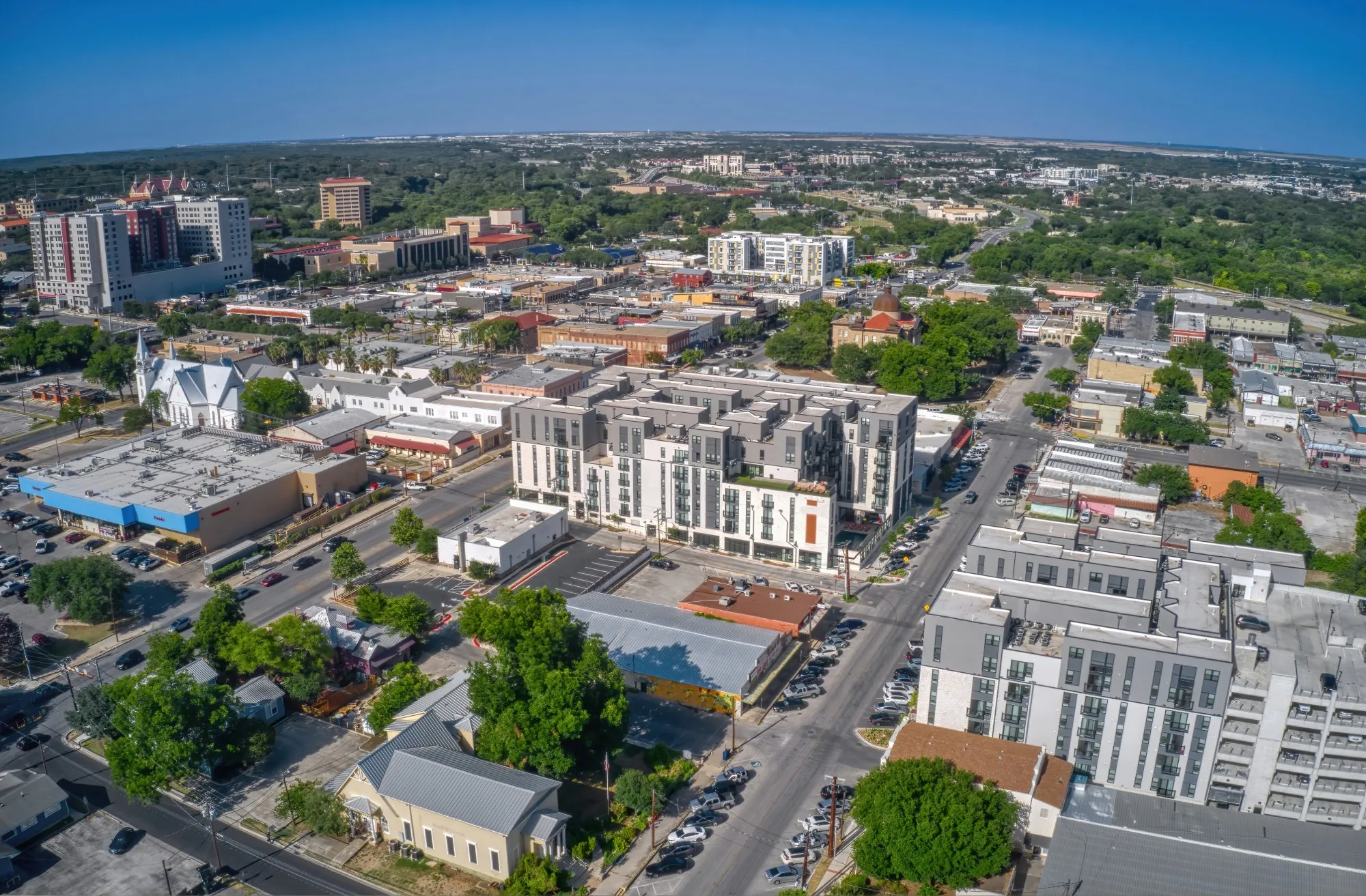 Aerial view of a cityscape featuring a mix of modern apartment buildings, commercial structures, and green tree coverage under a clear blue sky.