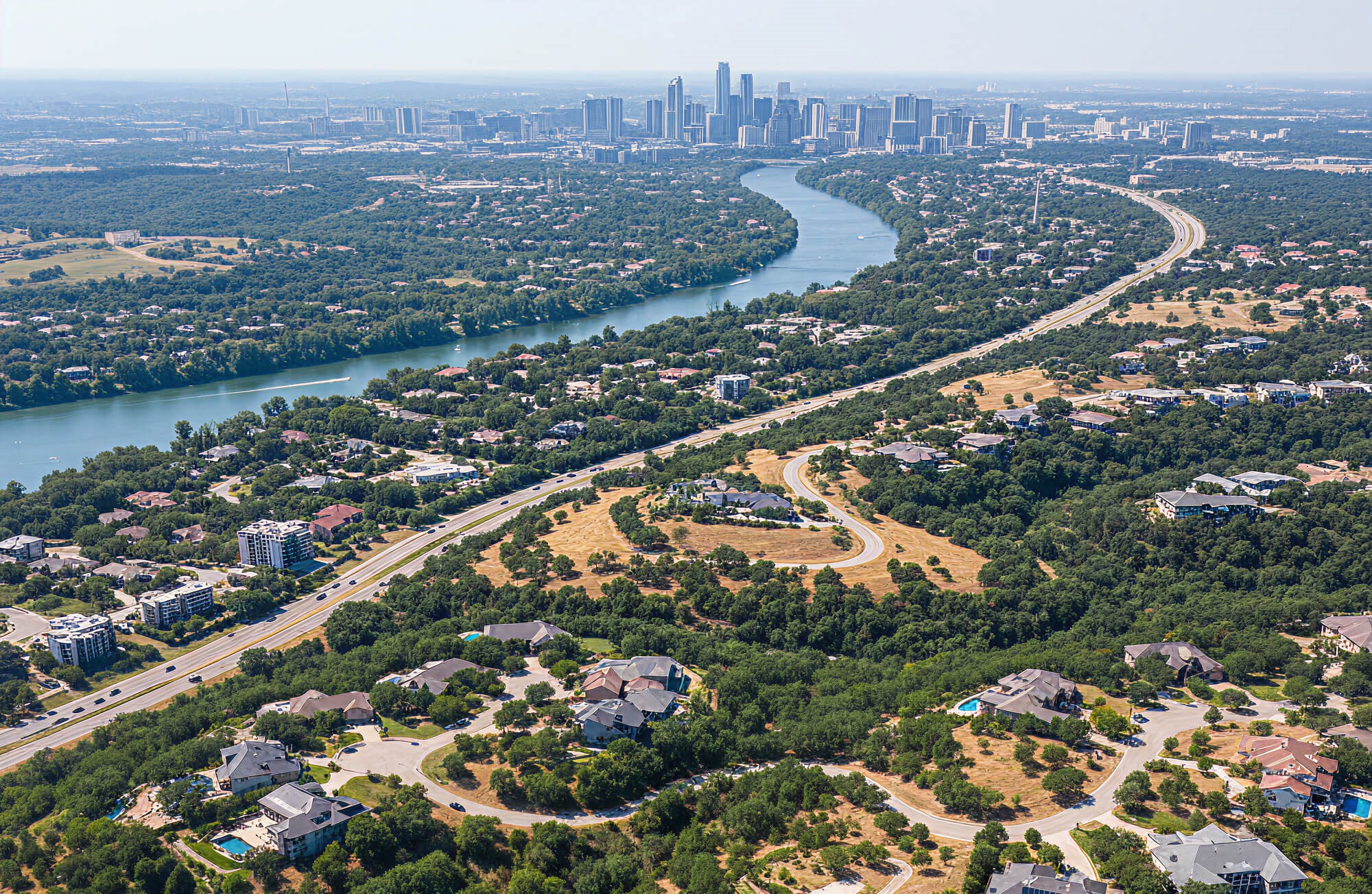 Aerial view of West Lake Hills, Texas, with the Colorado River winding through tree-lined residential neighborhoods and a highway, while the Austin skyline appears in the background. Fox Granite serves this area.