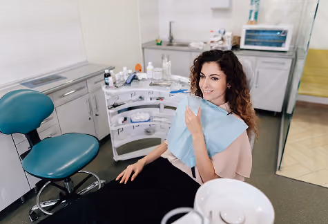 A woman with curly hair seated in a dental clinic chair smiling and holding a blue dental bib, with dental equipment and cabinets in the background.