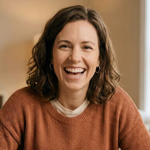 Smiling woman with curly brown hair wearing a rust-colored sweater and hoop earrings against a blurred indoor background.