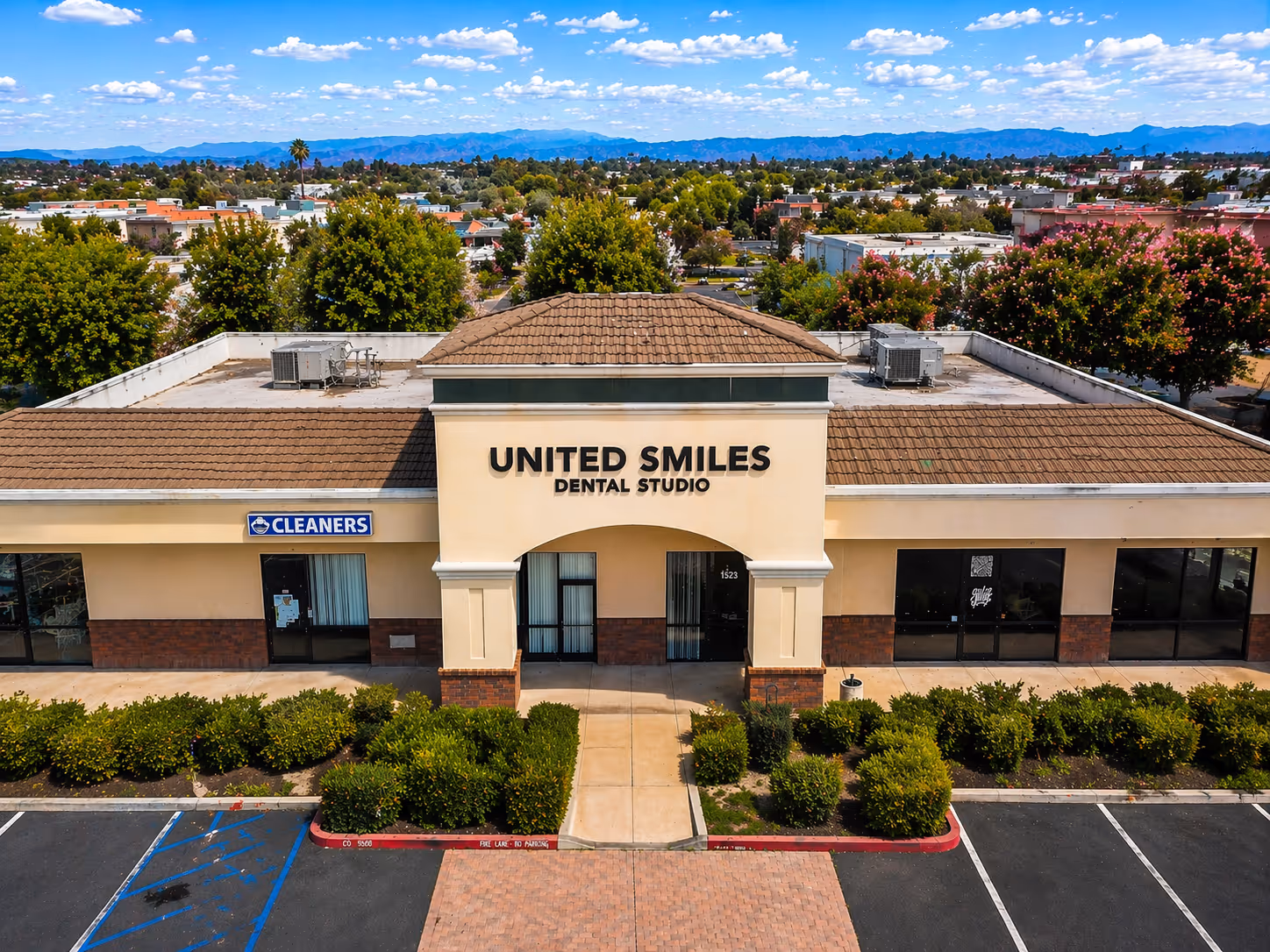 Front view of a commercial building with United Smiles Dental Studio in the center and a Cleaners shop on the left, surrounded by shrubs and parking spaces under a partly cloudy sky.
