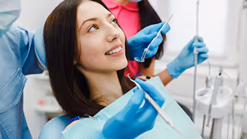 Young woman sitting in a dental chair smiling while a dentist wearing blue gloves holds dental tools near her mouth.