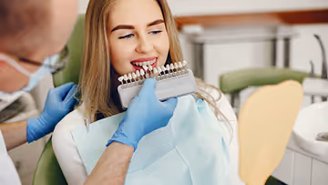 Dentist wearing blue gloves holding a shade guide next to a smiling woman's teeth to match dental implant color.
