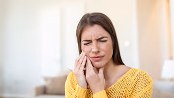 Woman in a yellow sweater pressing her fingers to her cheek with a pained expression, indicating toothache or facial pain.