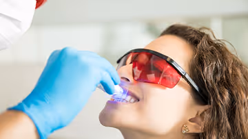 A woman wearing protective red-tinted glasses smiling as a dental professional wearing blue gloves applies a blue light device to her upper teeth during a teeth whitening procedure.
