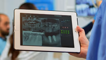 Hand holding a tablet displaying a digital dental X-ray image, with two blurred individuals in the background in a clinical setting.