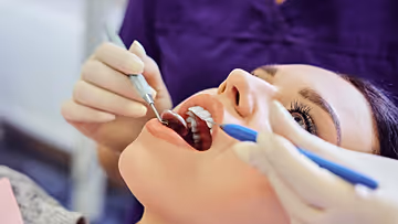 A dental professional wearing gloves uses dental instruments to examine a smiling woman's teeth during a dental checkup.