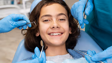 Smiling young girl reclined in a dental chair while a dentist wearing blue gloves holds dental tools near her face.
