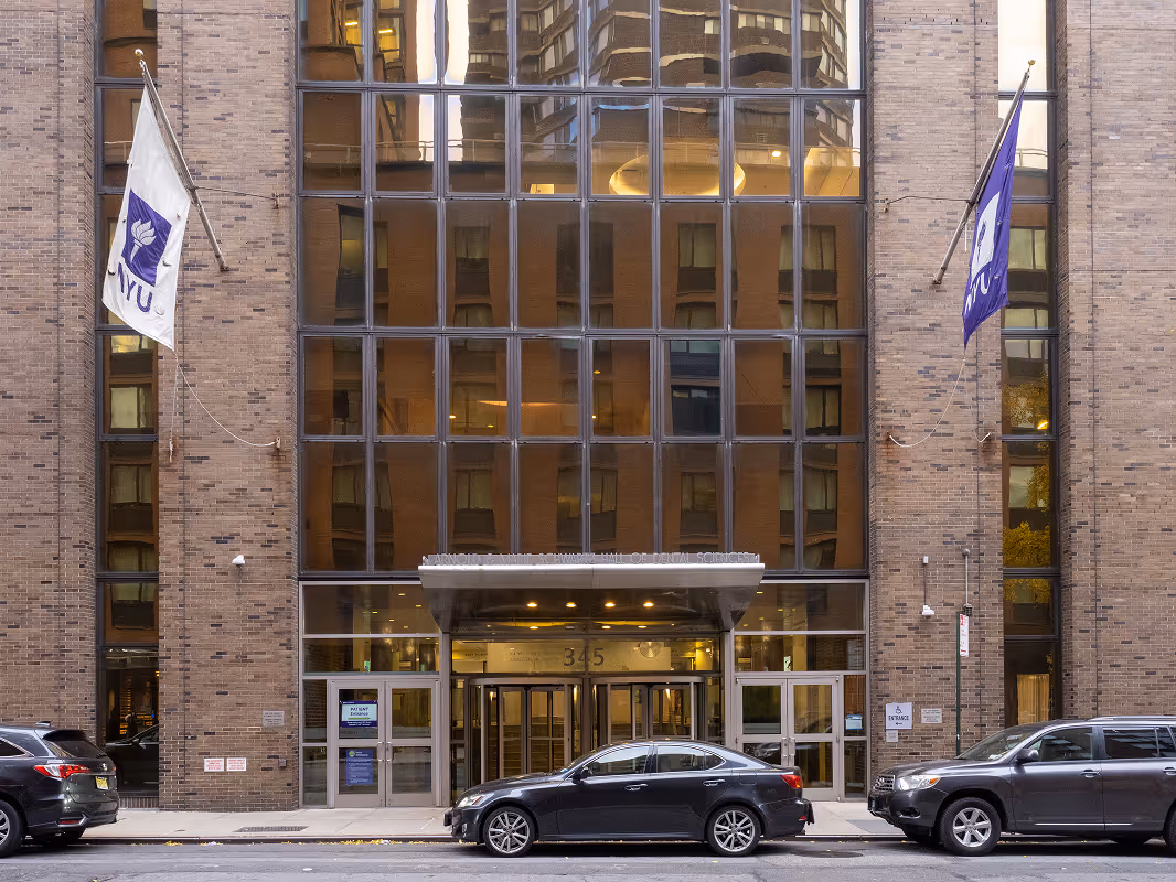 Exterior view of a building entrance with large glass windows, two NYU flags on either side, and three parked cars in front.