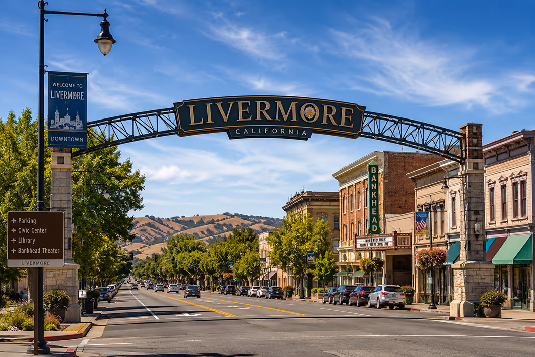 Street view of downtown Livermore, California, featuring an arch with 'LIVERMORE CALIFORNIA' over the road, historic buildings, parked cars, and a clear blue sky with light clouds.