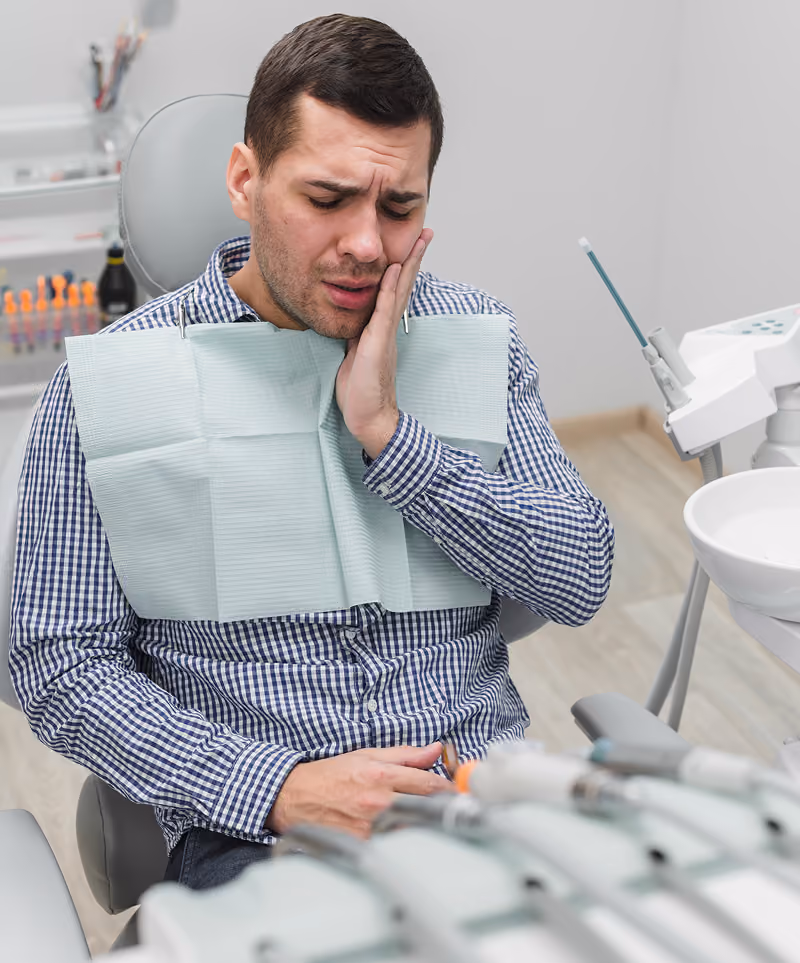 Man sitting in a dental chair wearing a dental bib, holding his cheek with a pained expression, indicating tooth pain or dental discomfort in a dentist's office.