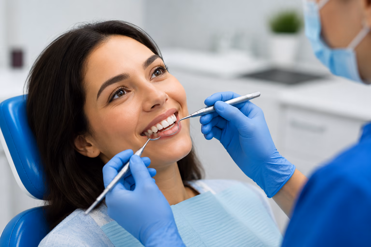 A woman reclining in a dental chair smiling while a dentist wearing blue gloves and a mask examines her teeth with dental tools in a clinical setting.