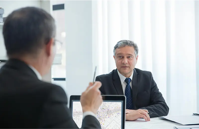 Zdjęcie ukazuje dwóch mężczyzn w garniturach rozmawiających na tematy biznesowe. Jeden z nich wyraźnie sugeruje drugiemu jakieś rozwiązanie.