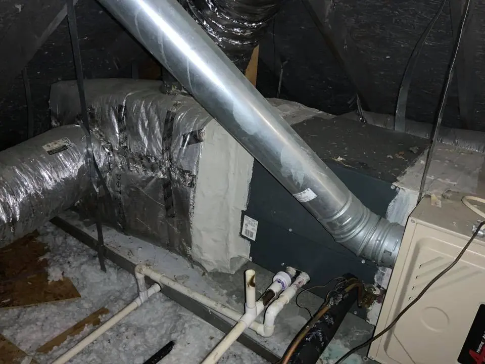 Furnace and ductwork installation in an attic with visible piping and insulation.