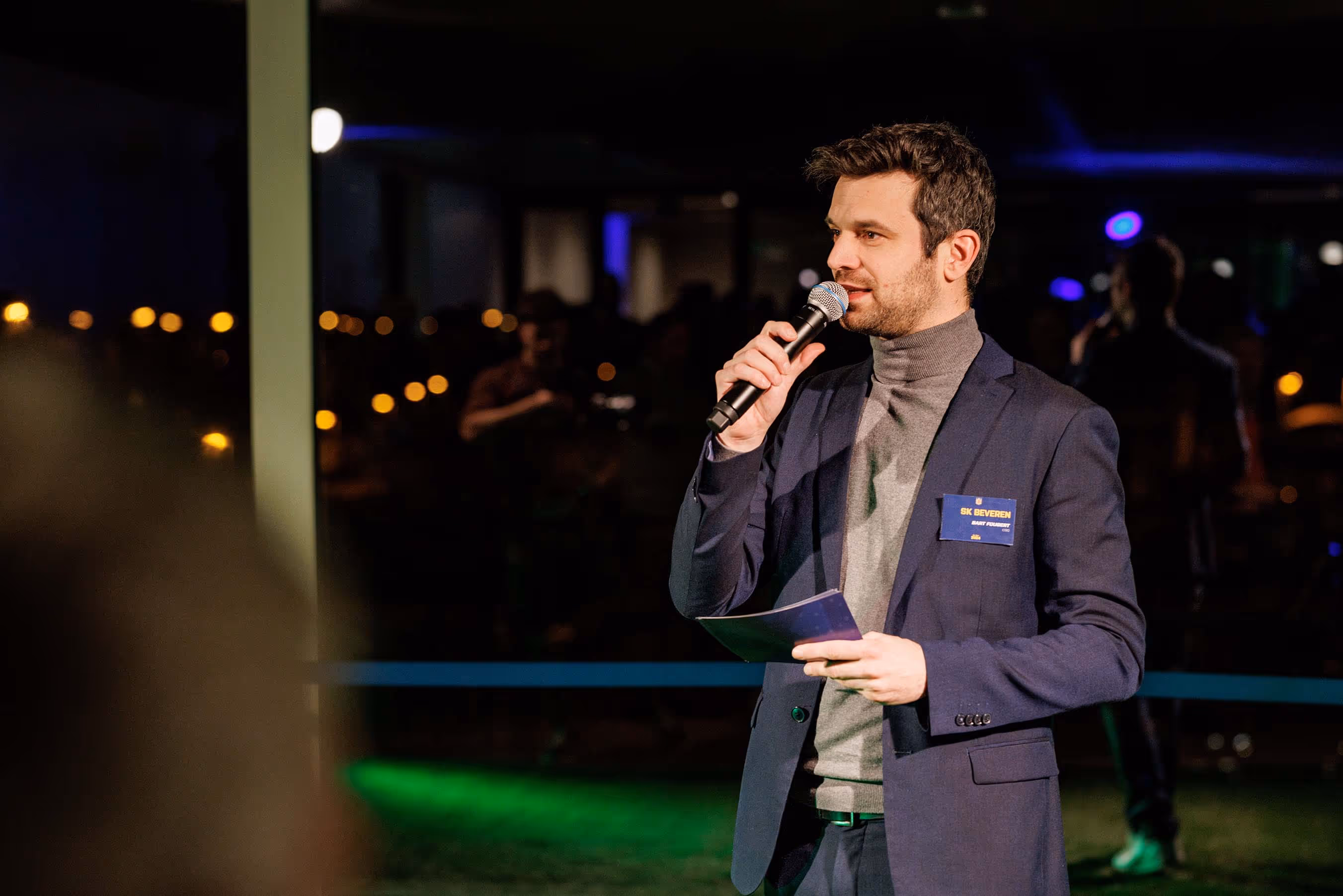 Man in a navy blazer and gray turtleneck speaking into a microphone while holding notes at an evening event.