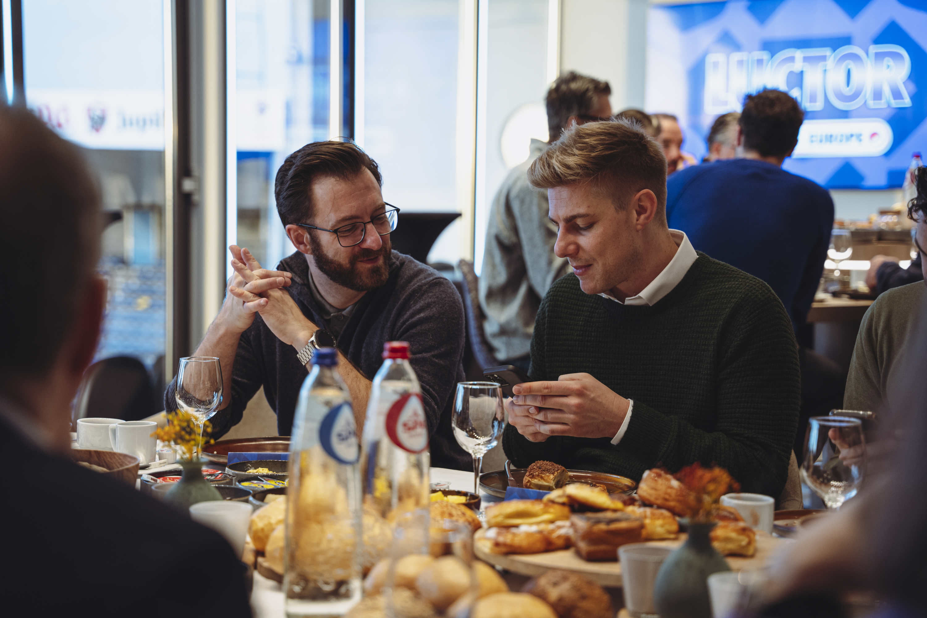 Two men sitting at a table with pastries and drinks, one looking at his phone and the other watching him.