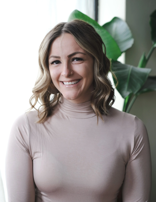 Sam Stroeh is a medical professional who assesses eligibility for KAP and psychiatric medication. Here she smiles with a plant behind her. She's wearing a light pink turtleneck