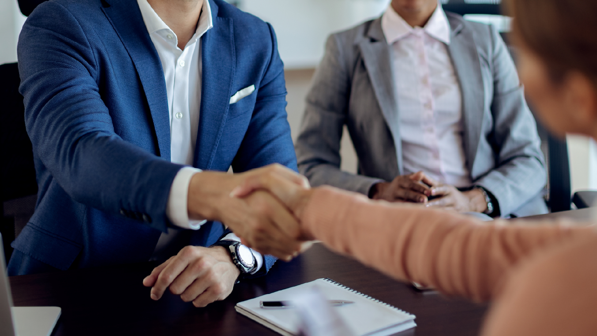 Two business professionals shaking hands across a table during a meeting, symbolising partnership and collaboration.