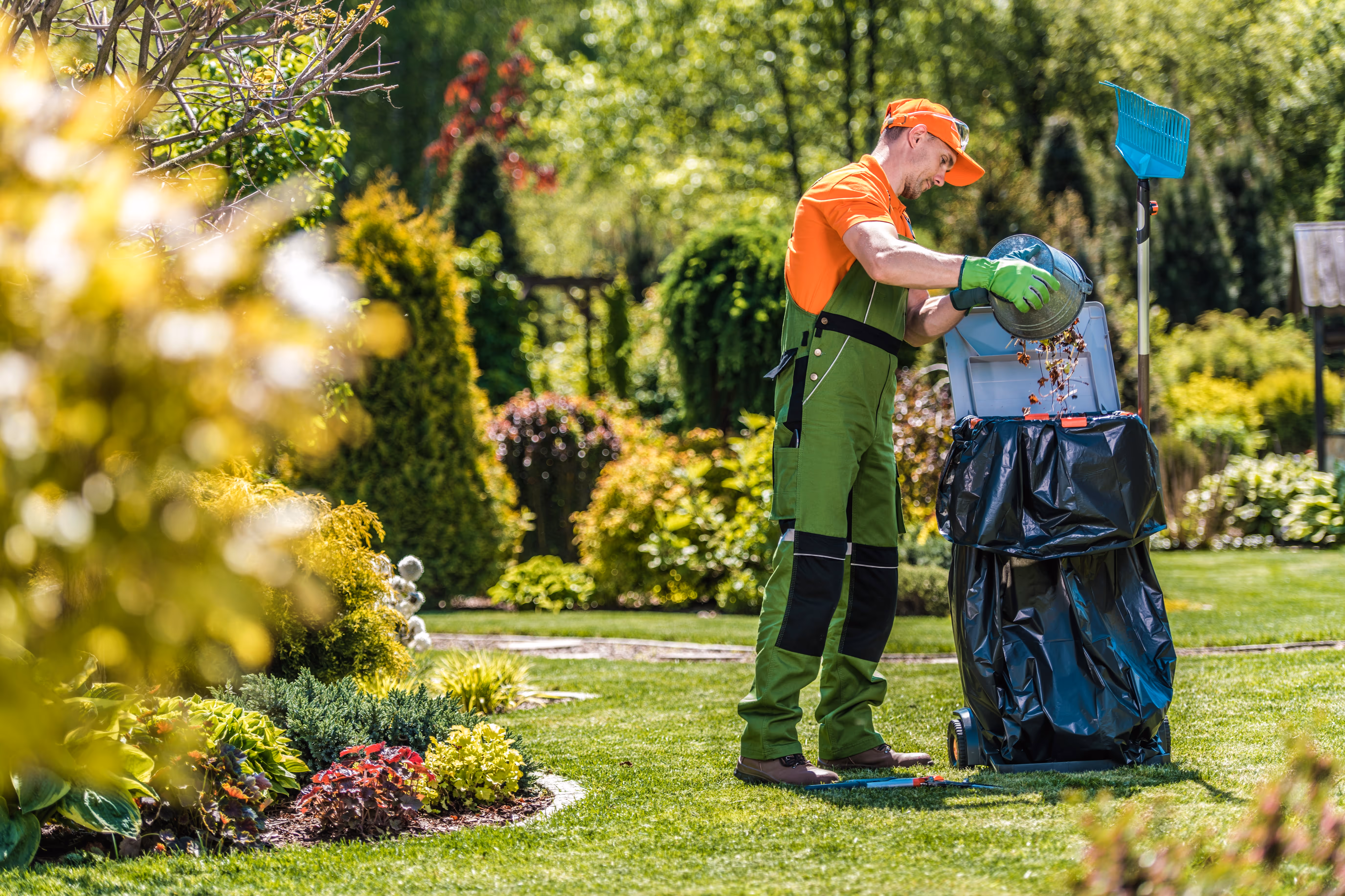 Worker performing a garden clean up, removing fallen leaves and debris with tools while surrounded by lush greenery.