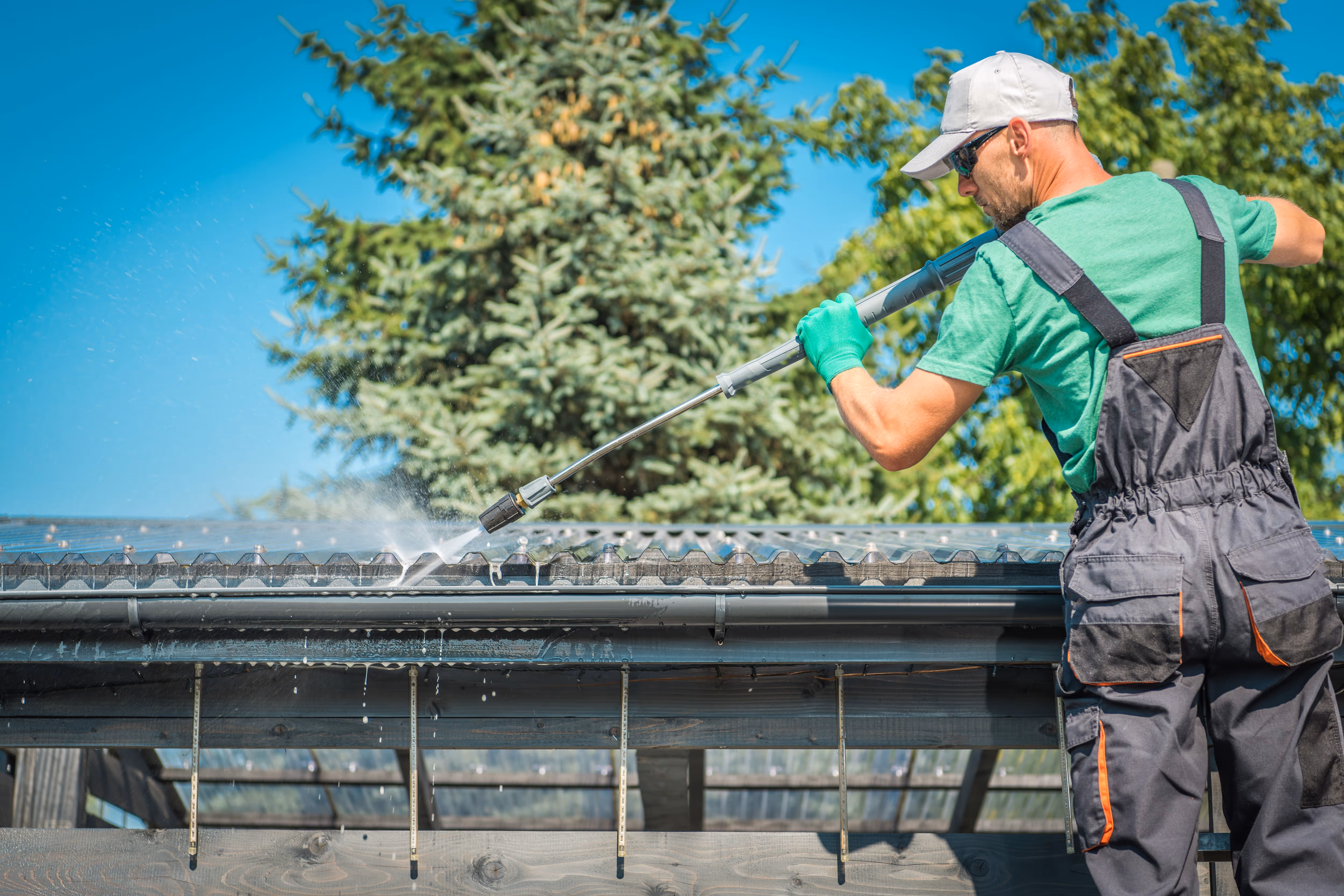 Worker in overalls and gloves using a pressure washer for gutter cleaning on a rooftop edge. Water sprays from the nozzle as debris is cleared, with trees and blue sky in the background highlighting outdoor maintenance work.
