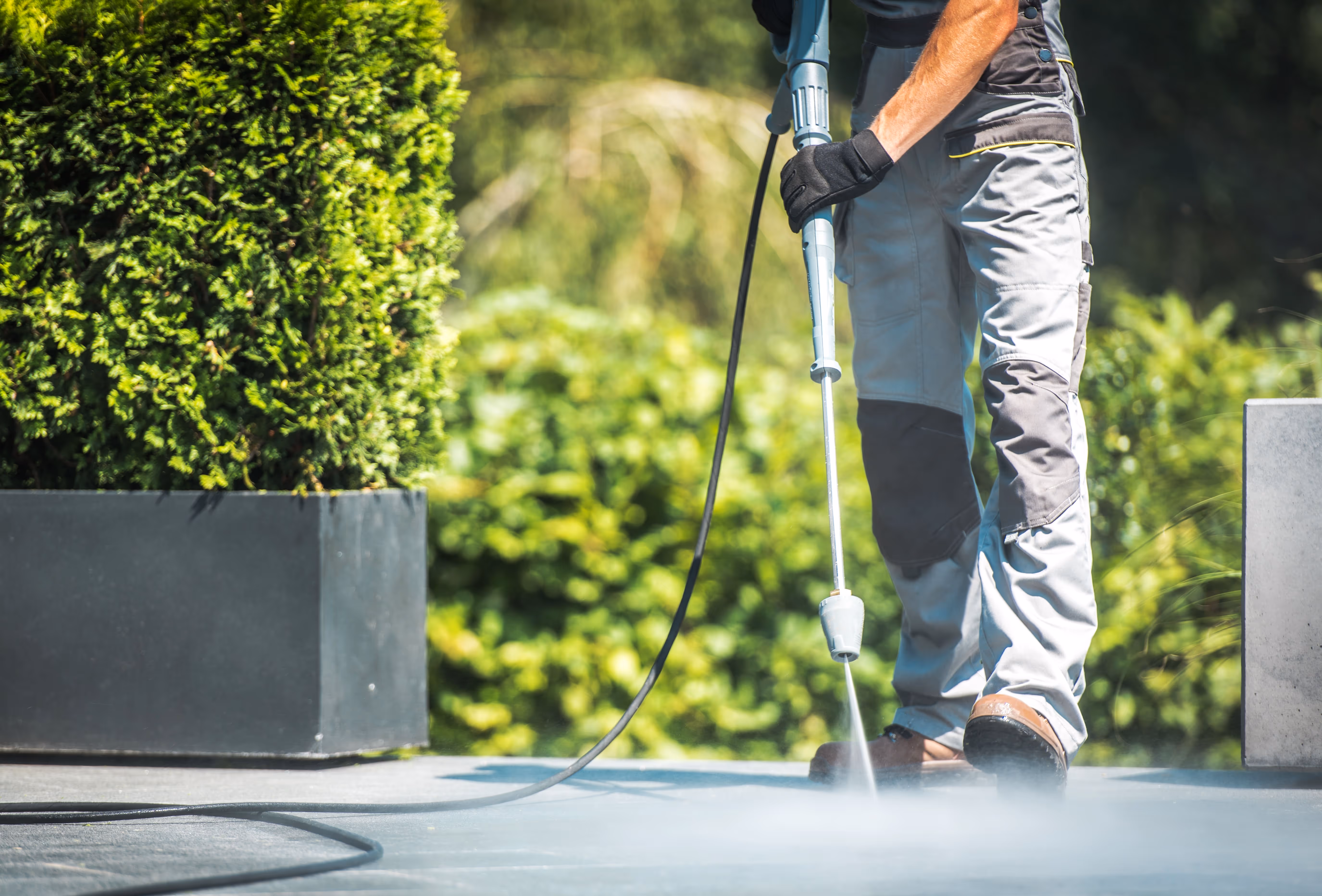 Worker wearing protective gloves and boots performing pressure washing on a concrete surface. A powerful stream of water sprays from the pressure washer, with greenery and potted plants in the background highlighting outdoor surface cleaning.