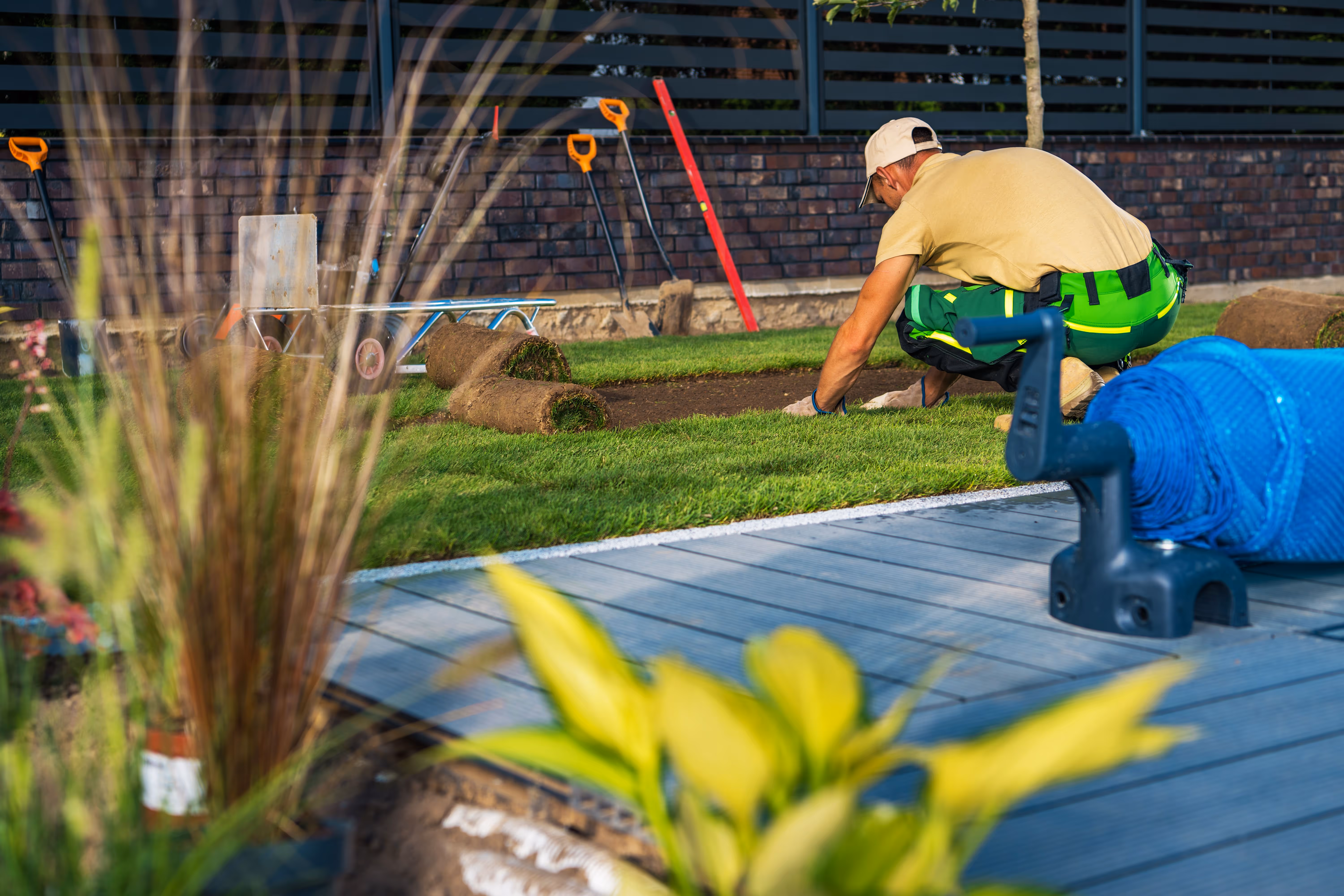 Landscaper in workwear laying rolled turf onto soil in a backyard garden, with tools and mulch bags nearby. The scene highlights a turf laying and mulching project, with fresh grass installation and landscaping materials in use.