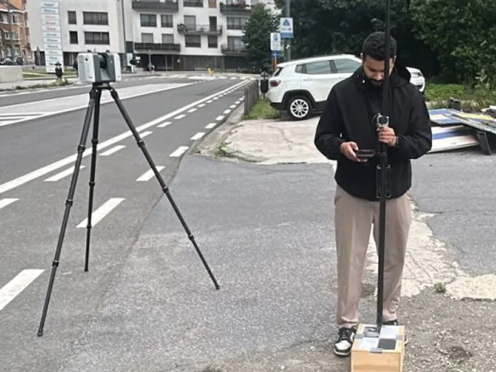 Géomètre relevant une voirie bruxelloise, récepteur GNSS en main, scanner laser sur trépied en bord de chaussée.