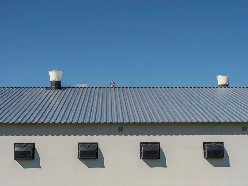 Szürke trapézlemez tető világoskék ég alatt, fehér falon négy fekete szellőzőnyílás.