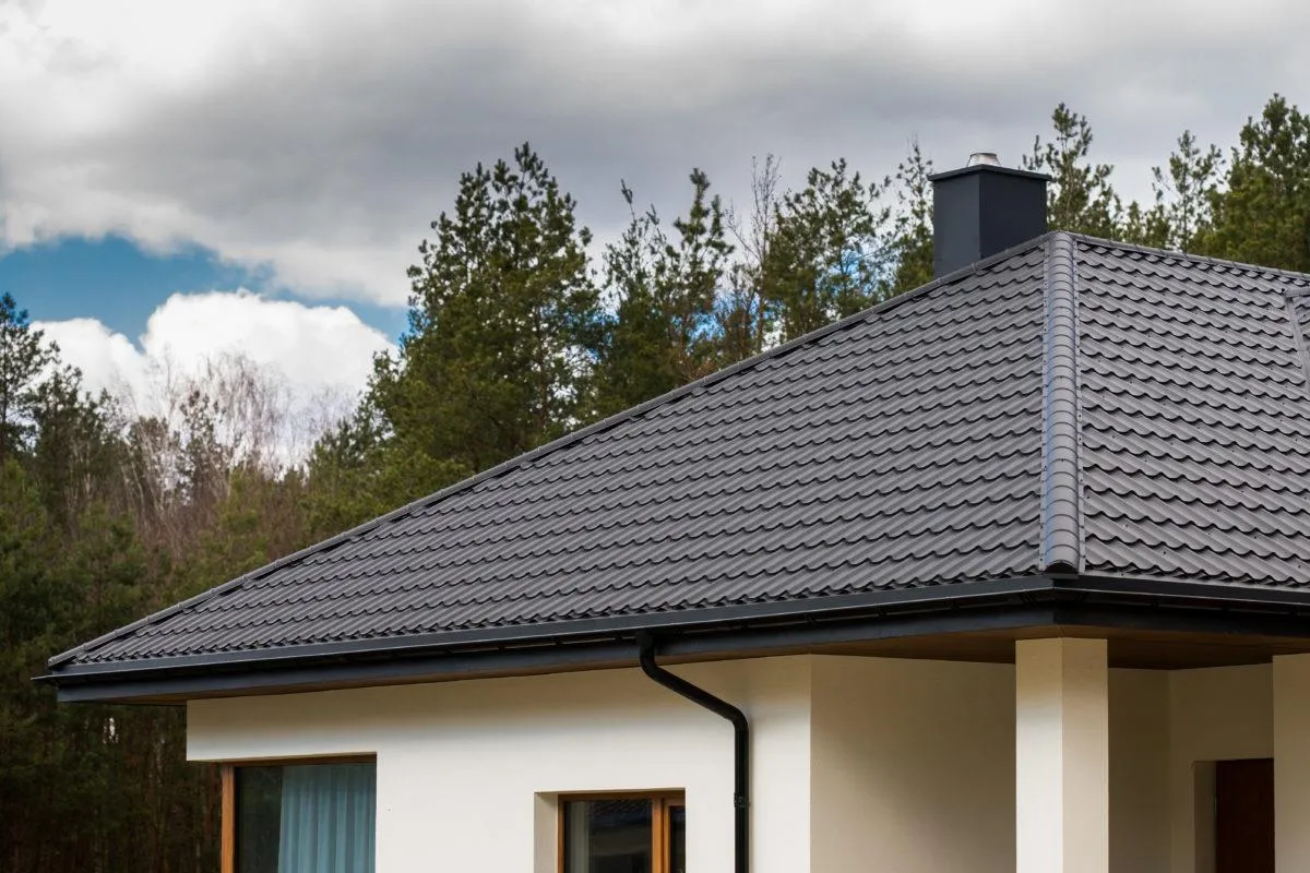 Egy modern ház sötétszürke hullámos tetővel és világos falakkal, háttérben fák és felhők.